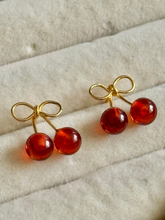 Cherry-shaped earrings with gold bow details on a textured beige surface