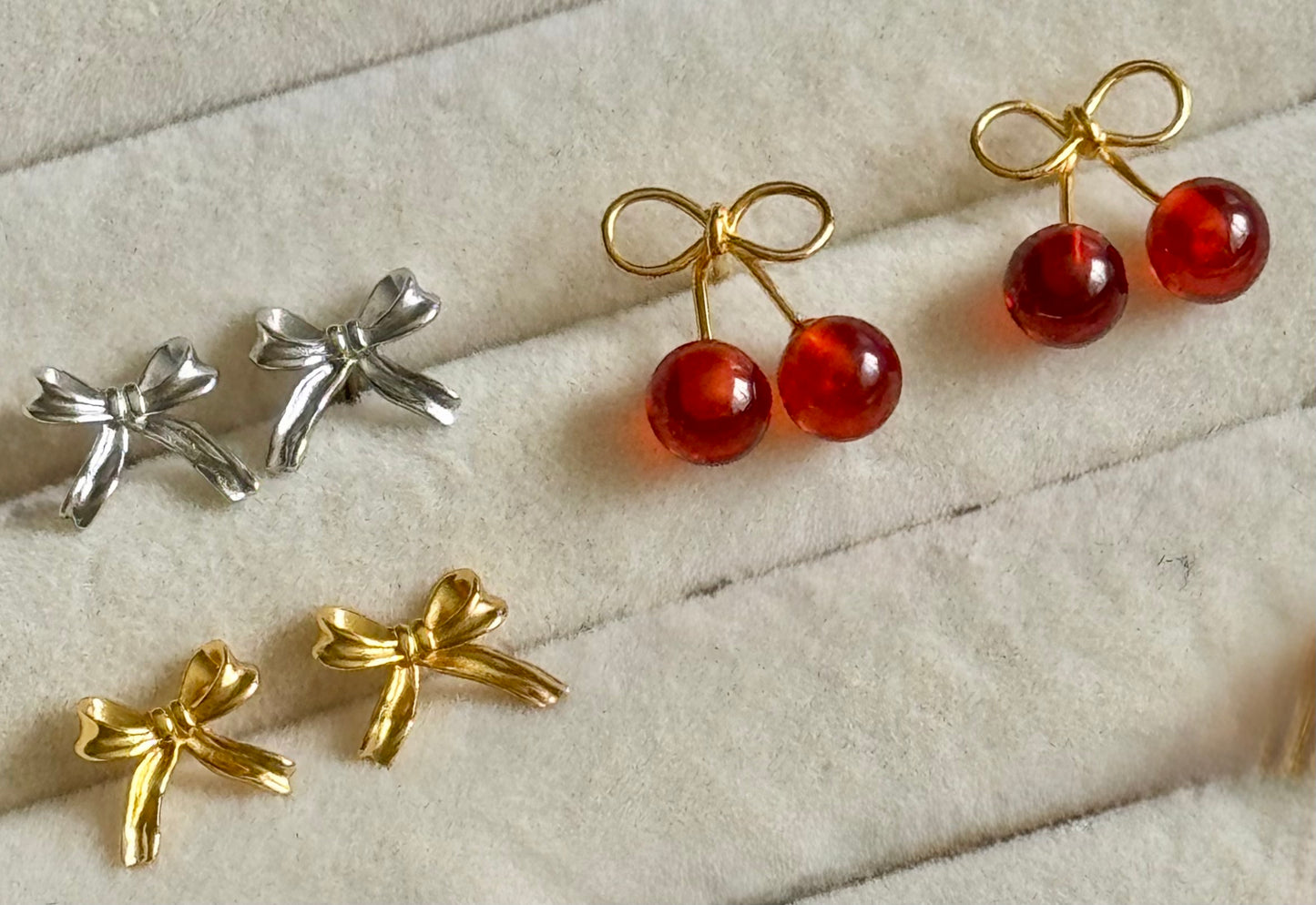 Pair of silver bow earrings and a pair of gold cherry earrings on a textured surface.